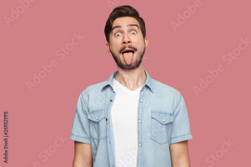 Portrait of funny crazy handsome bearded young man in blue casual style shirt standing with big eyes, tongue out and looking at camera. indoor studio shot, isolated on pink background.