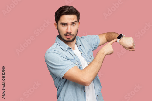 Time is out. Portrait of serious handsome bearded young man in blue casual style shirt standing and looking at camera, pointing on his smart watch. indoor studio shot, isolated on pink background.
