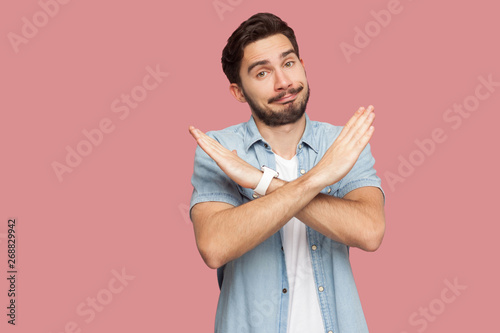 Portrait of serious handsome bearded young man in blue casual style shirt standing with X sign hands and looking at camera. indoor studio shot, isolated on pink background. photo