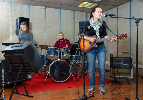 Attractive female soloist playing guitar and singing with her music band in sound studio
