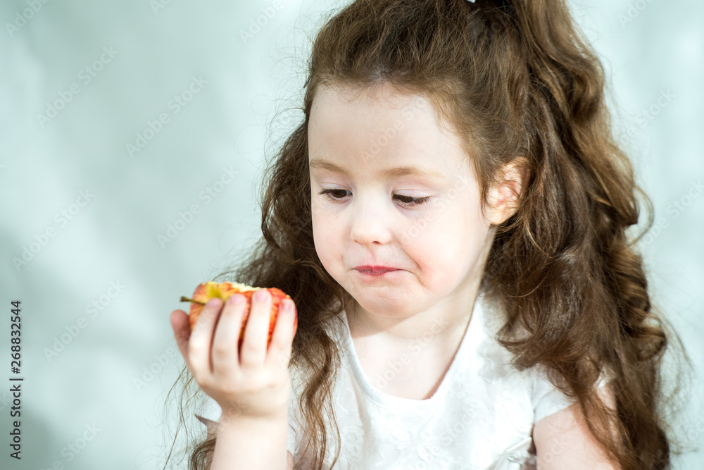 Cute little girl nibbles an apple with pleasure. Sun glare on the face. Emotional portrait of a little beautiful girl holding an apple. Studio 4-5 years. Cute little girl nibbles an apple with pleasure. Sun glare on the face. Emotional portrait of a little beautiful girl holding an apple. Studio 4-5 years.