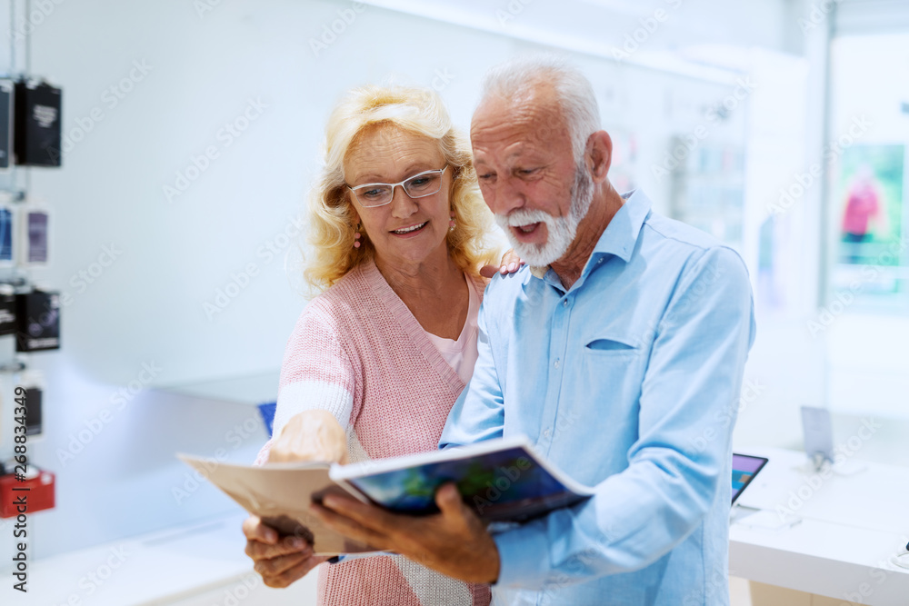 Fototapeta premium Cute smiling Caucasian senior couple looking at catalog in tech store.