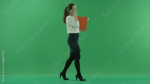 business woman is talking on her mobile cell phone and walking with folders. She wears formal dress: white shirt and blak skirt. Businesswoman is over green screen