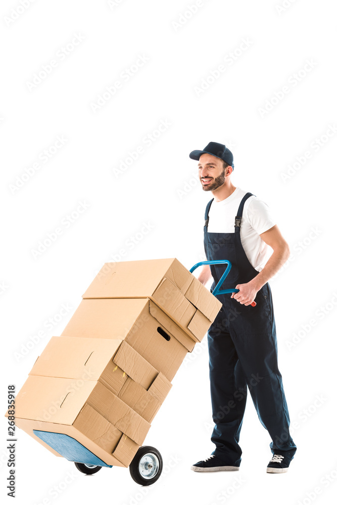cheerful handsome delivery man transporting cardboard boxes loaded on hand truck isolated on white cheerful handsome delivery man transporting cardboard boxes loaded on hand truck isolated on white