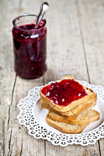 Toasted cereal bread slices on white plate and jar with homemade cherry jam closeup on rustic wooden table background.