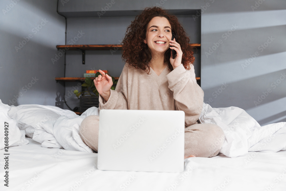 Image of beautiful young woman using laptop and cellphone while sitting in bed on white linen at home Image of beautiful young woman using laptop and cellphone while sitting in bed on white linen at home