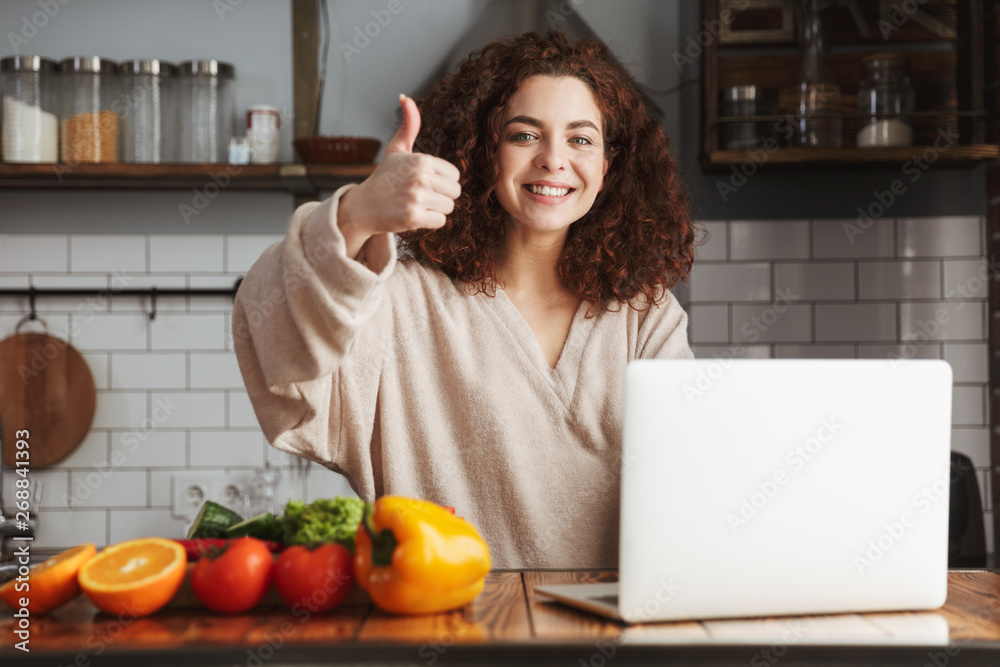 Image of young caucasian woman using laptop while cooking in kitchen interior at home Image of young caucasian woman using laptop while cooking in kitchen interior at home