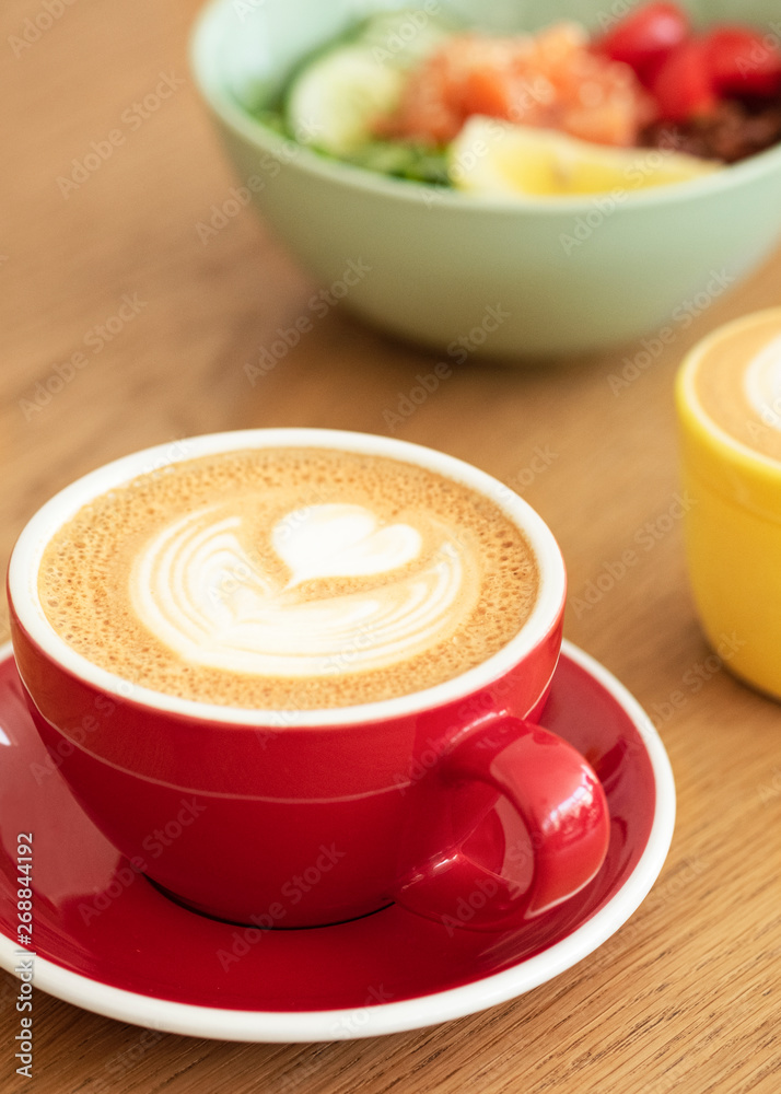 A red cup of latte with trendy latte art on a saucer with a coffee spoon. In the background a healthy breakfast bowl. A red cup of latte with trendy latte art on a saucer with a coffee spoon. In the background a healthy breakfast bowl.