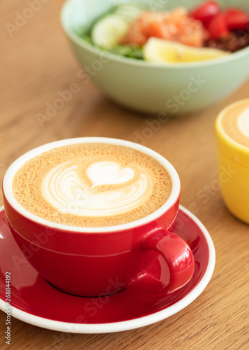 A red cup of latte with trendy latte art on a saucer with a coffee spoon. In the background a healthy breakfast bowl. 
