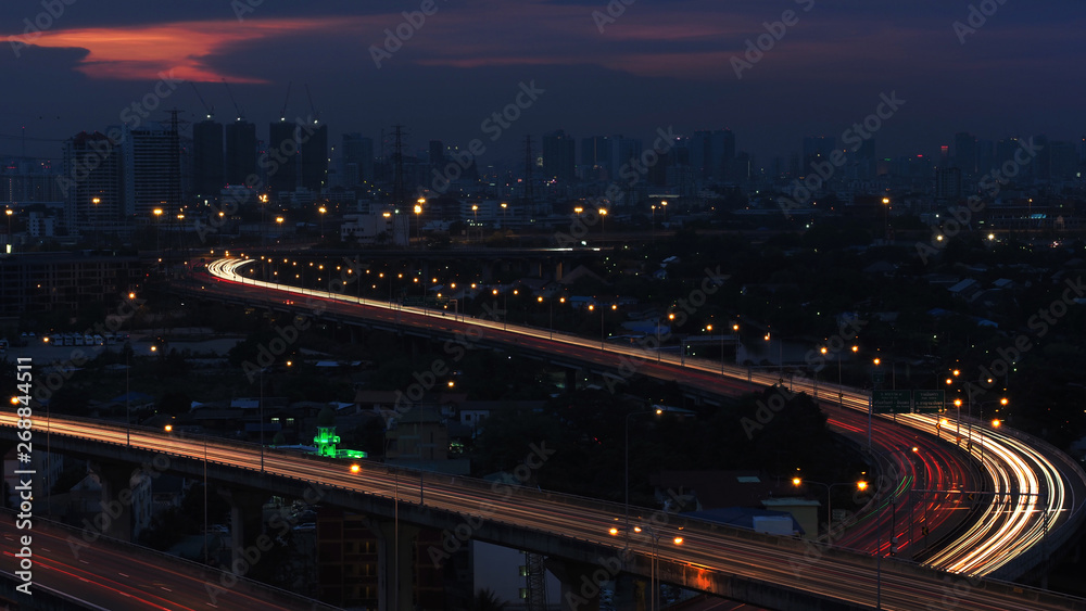 Motion blur of car light in city highway at dawn. Cross intersection of urban highways. Background have silhouette of cityand sunset purple orange sky.