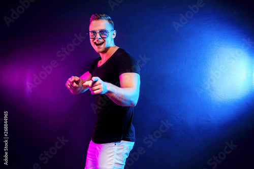 Excited young male in trendy outfit pointing at camera while having fun under colorful illumination during party