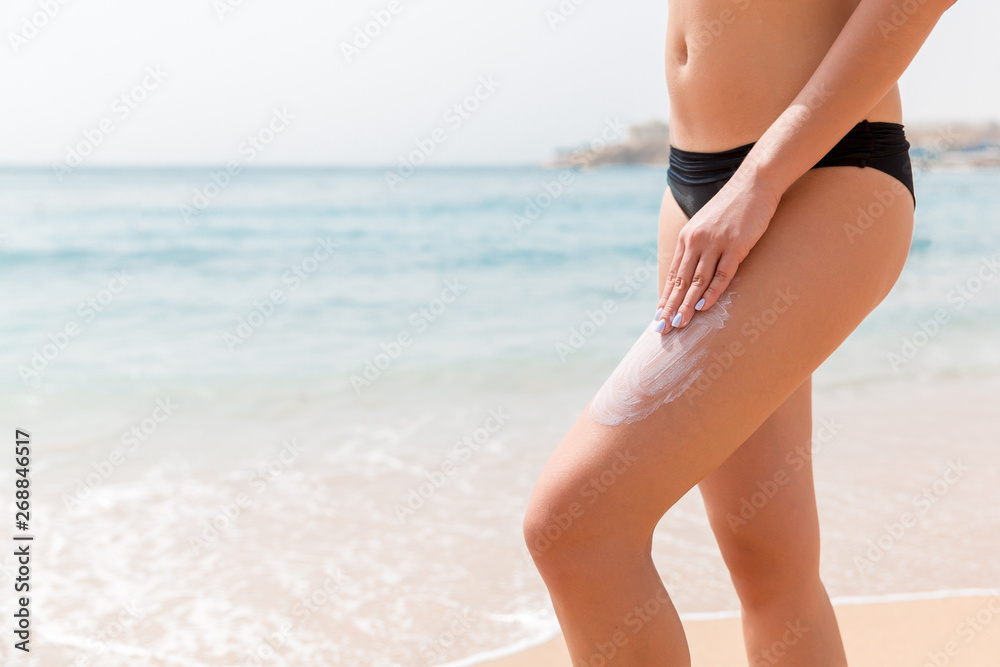 Woman in a black swimsuit is applying sun cream with her fingers on her leg at the sea background Woman in a black swimsuit is applying sun cream with her fingers on her leg at the sea background