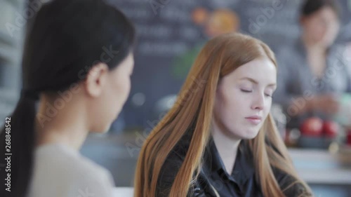 Wallpaper Mural Tracking right shot of two young females talking at table in cafe. Pretty red-haired young woman telling something to defocused Asian friend Torontodigital.ca