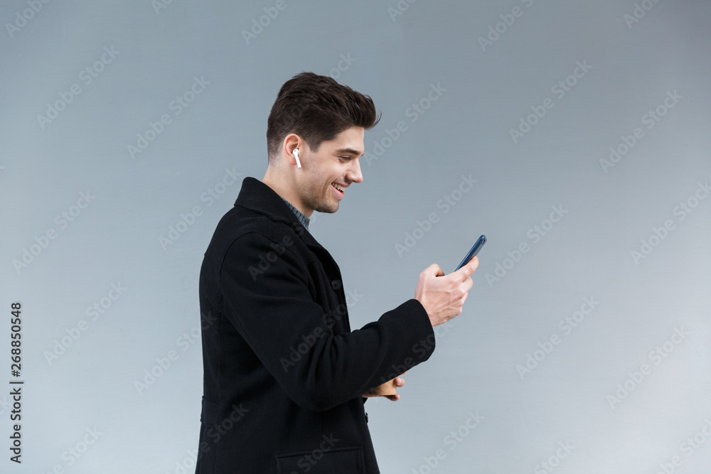 Image of cheerful young man wearing earpods holding takeaway coffee and mobile phone Image of cheerful young man wearing earpods holding takeaway coffee and mobile phone