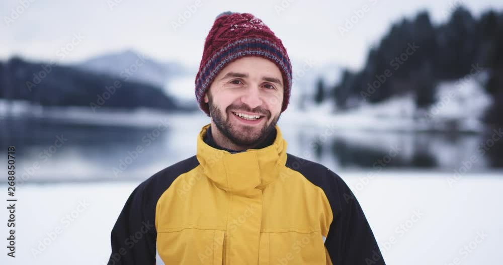 Closeup funny tourist smiling large in front of the camera , he wearing a red hat standing beside of lake in amazing place with snowy mountain and forest Closeup funny tourist smiling large in front of the camera , he wearing a red hat standing beside of lake in amazing place with snowy mountain and forest