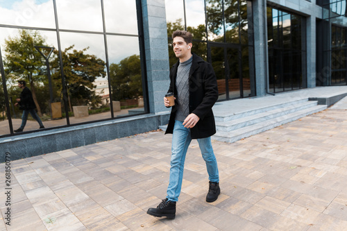 Portrait of cheerful man holding paper cup of takeaway coffee while strolling in front of glass building