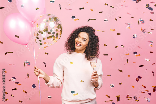 Party time! Happy beautiful african-american woman with glass of champagne, balloons and falling confetti isolated over pink background.