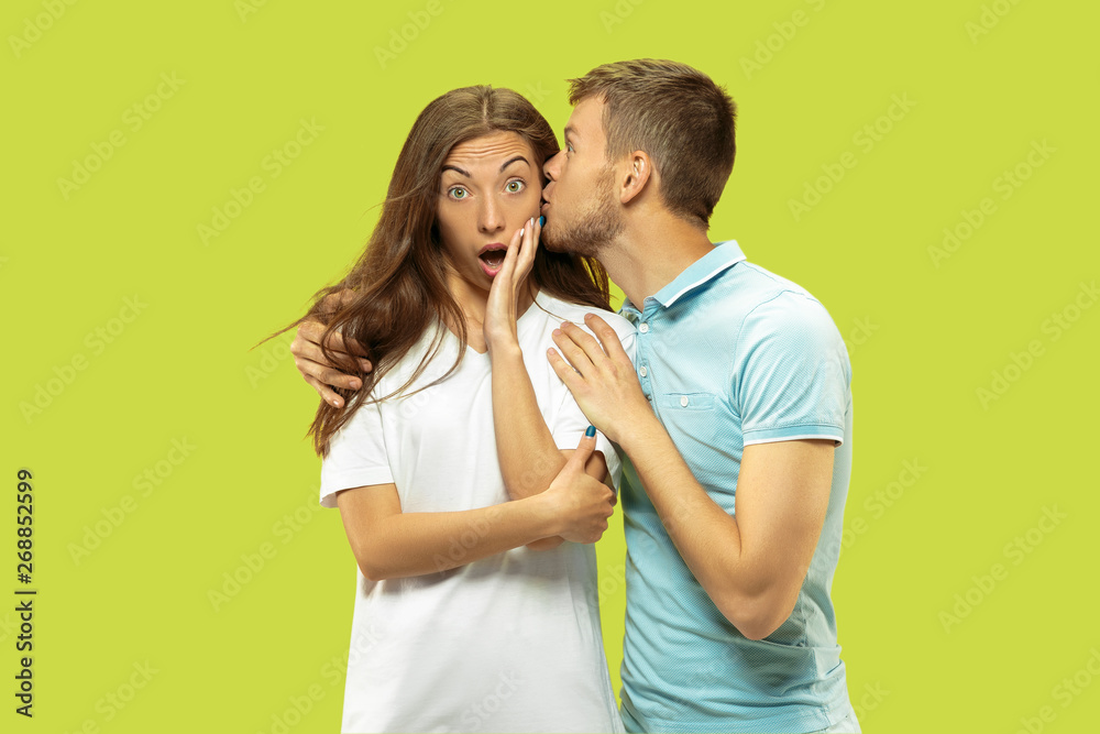 Naklejka premium Beautiful young couple's half-length portrait isolated on green studio background. Woman and man standing, caddling and kissing . Facial expression, human emotions concept. Trendy colors.