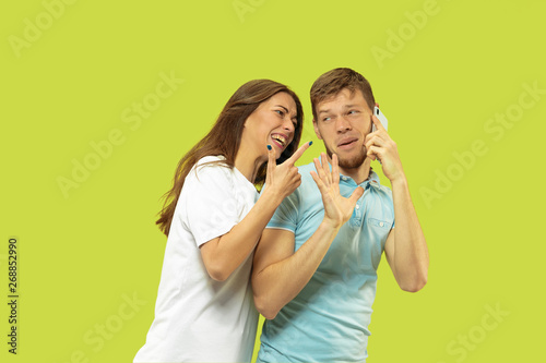 Beautiful young couple's half-length portrait isolated on green studio background. Man speaking on the phone, woman is angry for it. Facial expression, human emotions concept. Trendy colors.