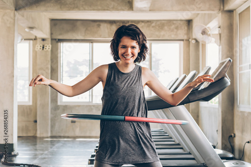 Beautiful young smiling happy Caucasian sporty woman with short hair playing hula hoop inside gym studio with treadmills behind - fun workout fitness portrait concept photo
