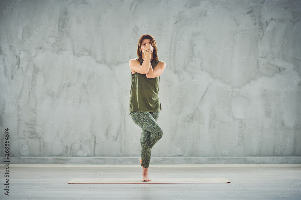 Fototapeta premium Full length of beautiful Caucasian brunette standing on the mat barefoot in Eagle yoga pose.