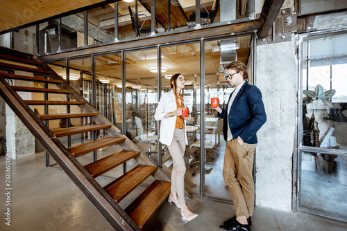 Young buiness man and woman having a conversation, sitting on the stairs during the coffee break in the modern office, wide interior view
