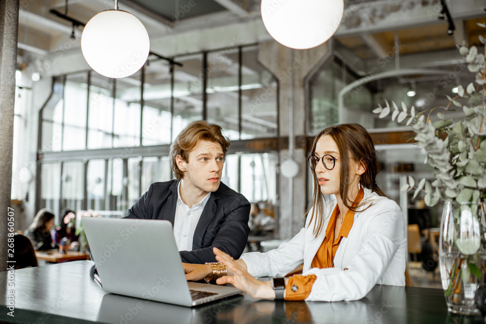 Young man and woman having a business conversation while sitting with laptop at the bar or modern spacious office Young man and woman having a business conversation while sitting with laptop at the bar or modern spacious office