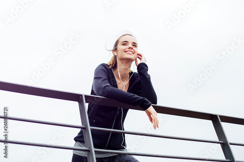 Sporty young woman with earphones leaning on fence and looking away outdoors