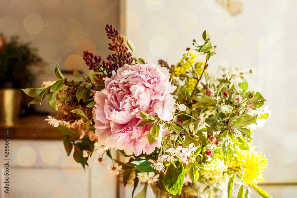 Fototapeta premium A spring bouquet of pink peony flowers, a blossoming apple tree branch, a lilac flower and wild flowers in a transparent jug are in the kitchen. Copy space.