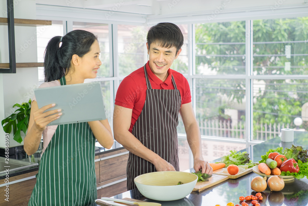 Asian couple are cooking in the kitchen. Man and Woman smiling looking menu from tablet. Man are cutting vegetables with knives. prepare salad food for diner. Asian couple are cooking in the kitchen. Man and Woman smiling looking menu from tablet. Man are cutting vegetables with knives. prepare salad food for diner.