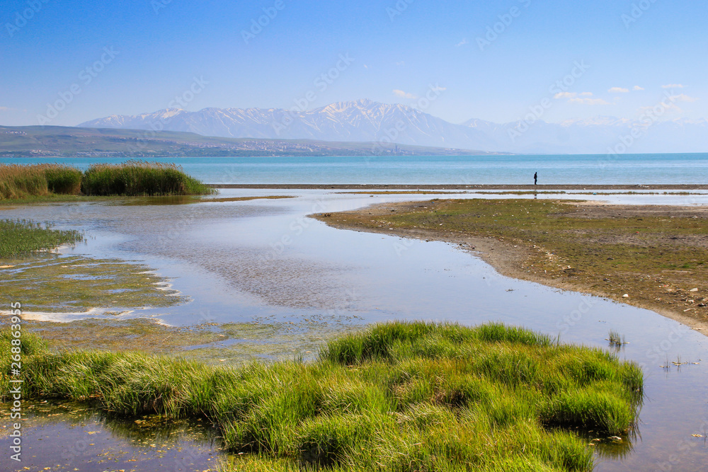 Van, Turkey - June 1, 2017: Wild beach with grass and bright clean turquoise water. Lonely male figure on a background of mountains.