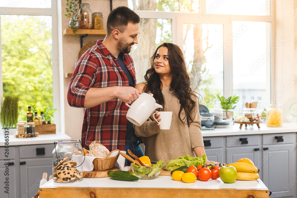 Beautiful young couple is talking, smiling while eating tea or coffee and drinking in kitchen at home. Beautiful young couple is talking, smiling while eating tea or coffee and drinking in kitchen at home.