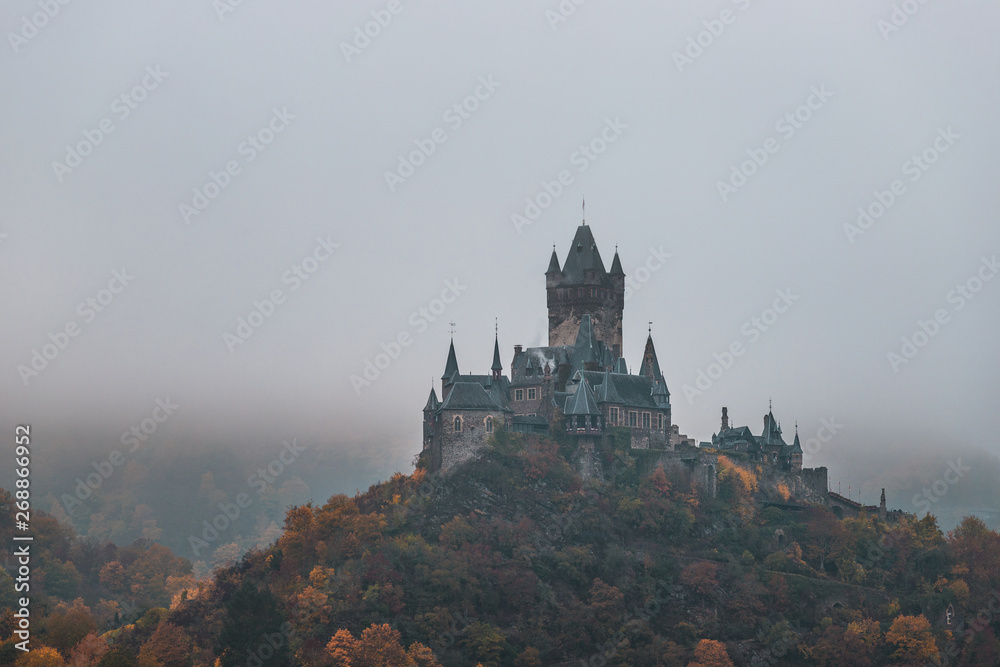 Reichsburg Cochem / Castle Cochem covered in fog during an orange and misty autumn day with low hanging clouds and fog (Cochem, Germany, Europe)
