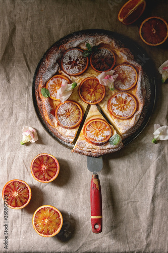 Cutted Homemade Cheesecake with sicilian blood oranges, decorated by edible flowers, mint leaves, sugar powder served in plate with cutted oranges above over grey linen table cloth. Flat lay, space