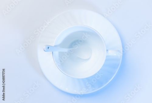 Empty white mug on a white saucer on a white table. Spoon in a mug. Top view