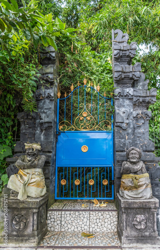 Bali, Indonesia - February 25, 2019: Ulun Danu Beratan Temple in Bedoegoel. Gray stone split gate or Candi Bentar gives access to green sanctuary. Blue metal port. Funny guard statues up front. photo