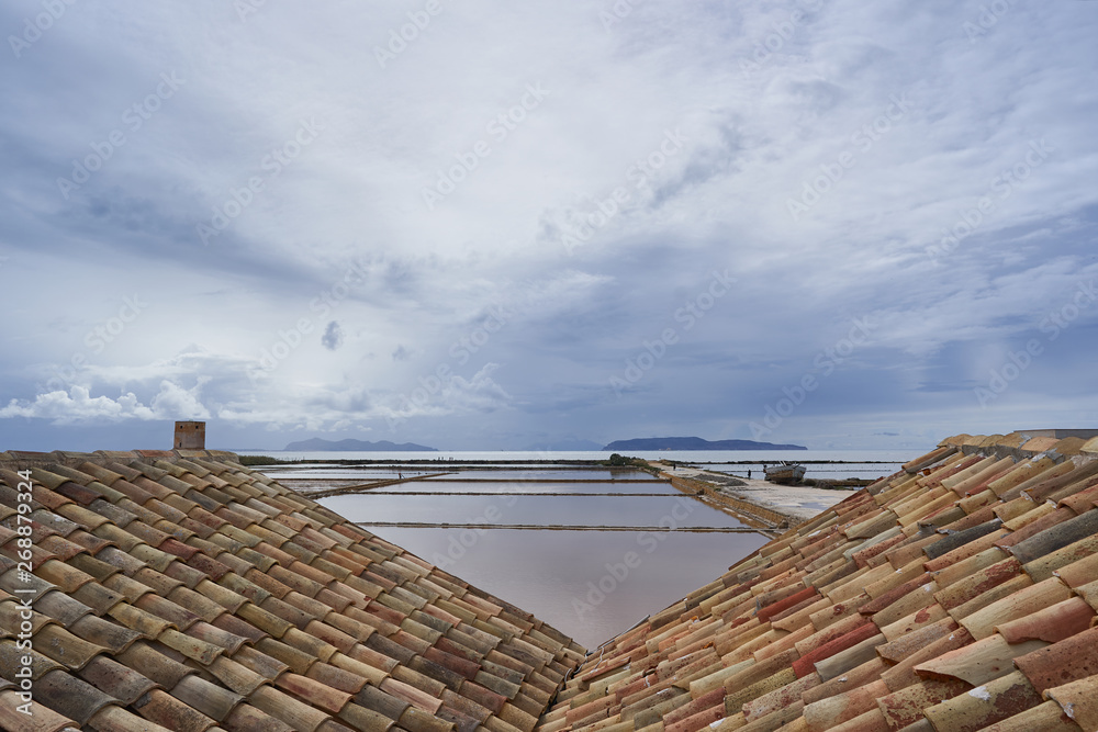 Evening landscape picture of ponds or saltpans in traditional salt production in Trapani in island Sicily during the sunset with cloudy sky and calm sea. Riserva naturale integrale Saline di Trapani.
