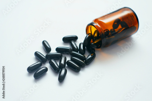 Black sleek capsules spill out from yellow glass bottle. White background. Medical concept.