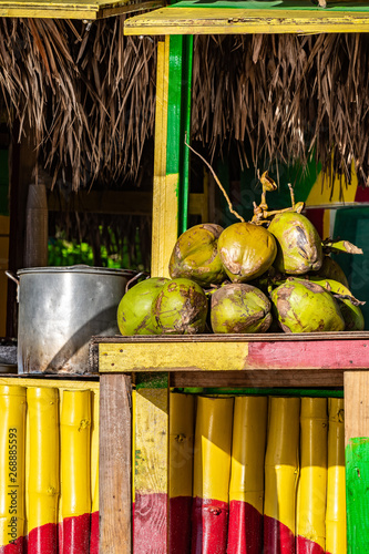 Tropical jelly coconut fruits on table at outdoor vendor shop painted in rasta colors with thatch roof. Big pot of Jamaican soup dish cooking in background. Sunny summer beach day setting in Jamaica.