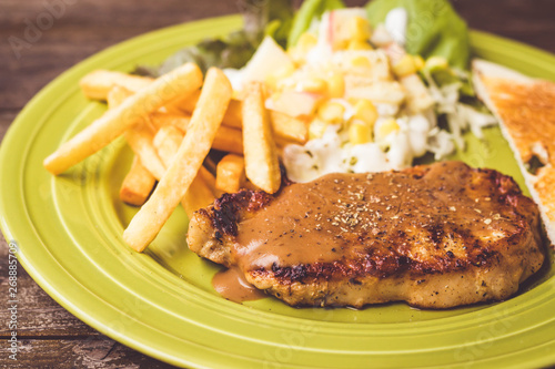 Pig steak is a fast food,pig steak this dish consists of with salad which has  purple lettuce corn apple cabbage salad dressing bread french fries and black pepper sauce, all in a green plate.