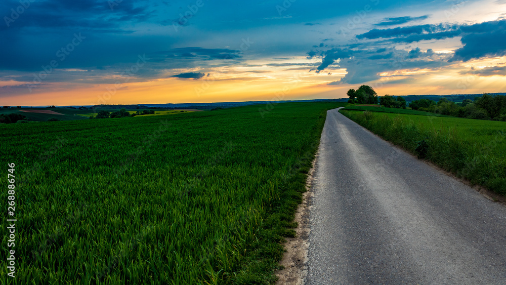 Empty country road in rural landscape against dramatic sunset sky at horizon; in Neudenau, Germany.