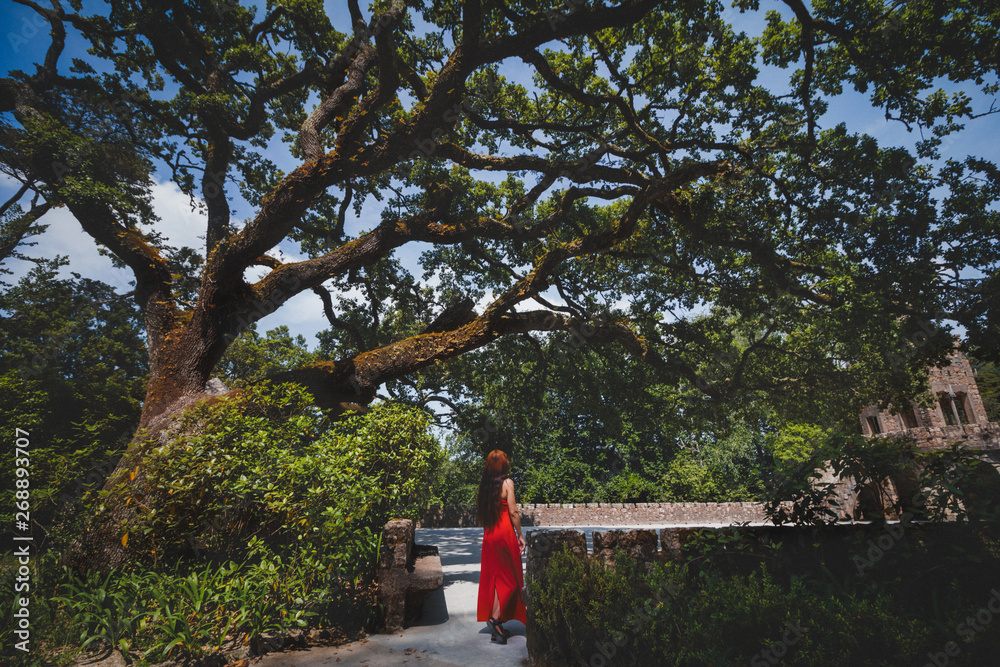 Romantic girl in a red dress is walking in the Regaleira Palace (Quinta da Regaleira) and a luxurious park located in Sintra. World Heritage Site by UNESCO. Principal tourist attraction of Portugal.