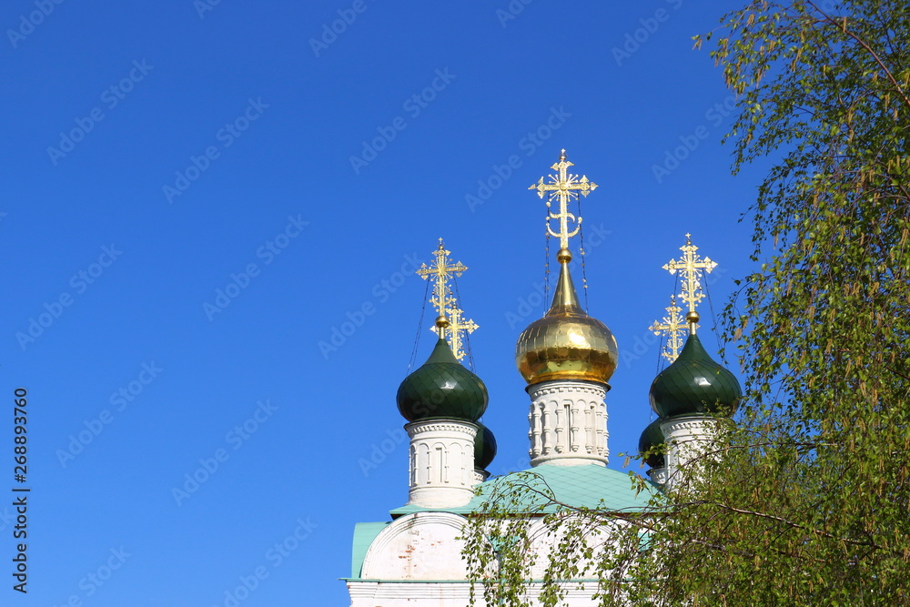Shined golden crosses on the green and golden orthodox domes of St. Nicholas Cathedral on blue sky background and green foliage birch. Background for religious greeting card with copy space