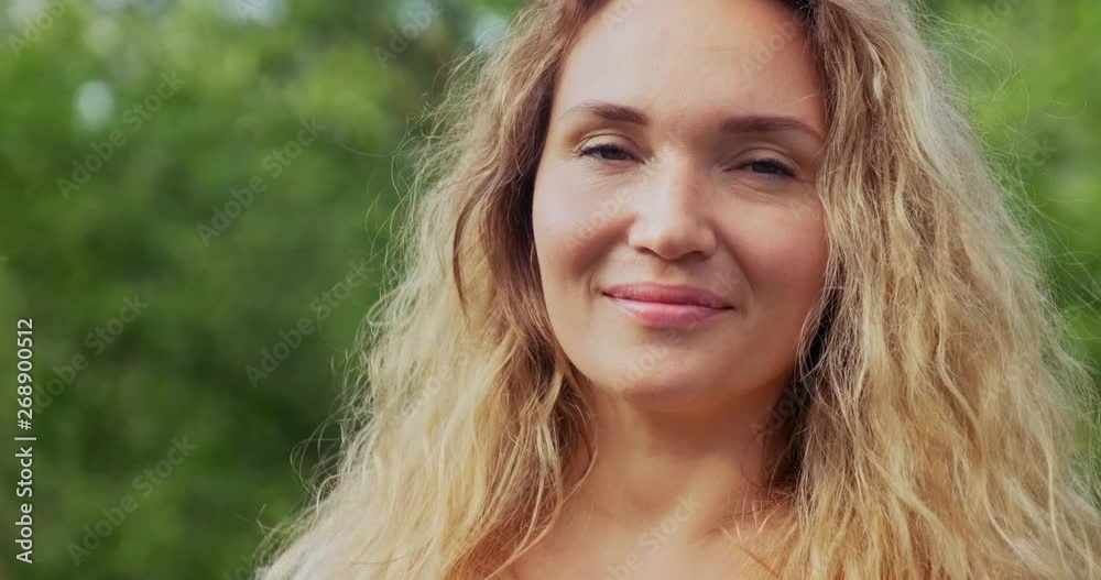 Closeup face of a happy adult woman on the nature looking at camera. Cheerful adult girl in the park. Outdoors portrait of a blonde woman with long curly hair in the park. Slow motion. Closeup face of a happy adult woman on the nature looking at camera. Cheerful adult girl in the park. Outdoors portrait of a blonde woman with long curly hair in the park. Slow motion.