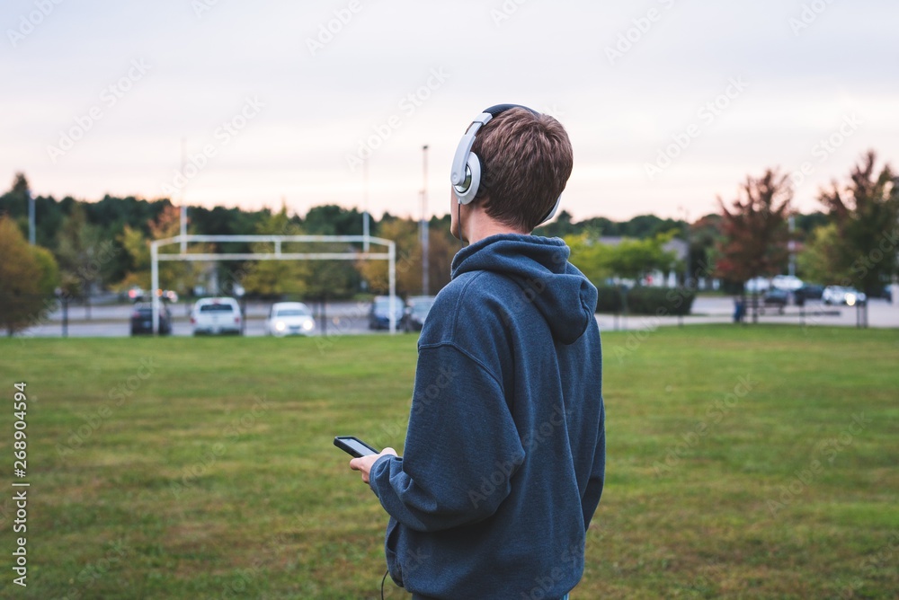 Teenager listening to music with his headphones. He is standing in the middle of a soccer field next to a school.