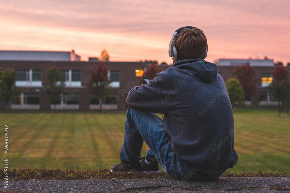 Teenager sitting alone at the top of a hill at sunset. He is listening to music through his headphones.