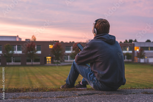 Teenager sitting alone at the top of a hill at sunset. He is listening to music through his headphones.