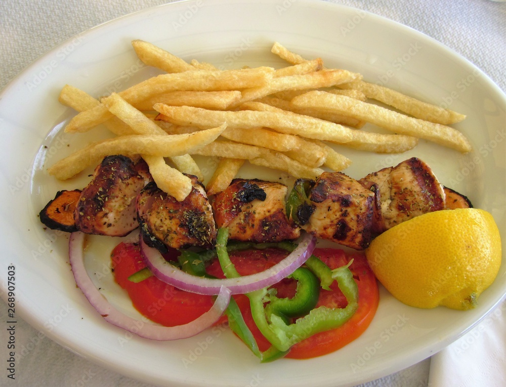 Chicken souvlaki with french fries and vegetables. White plate. Delicious greek traditional food. Chicken souvlaki with french fries and vegetables. White plate. Delicious greek traditional food.