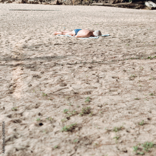 A lonely man lying on stomach sunbathing on the beach wearing a blue swimwear.