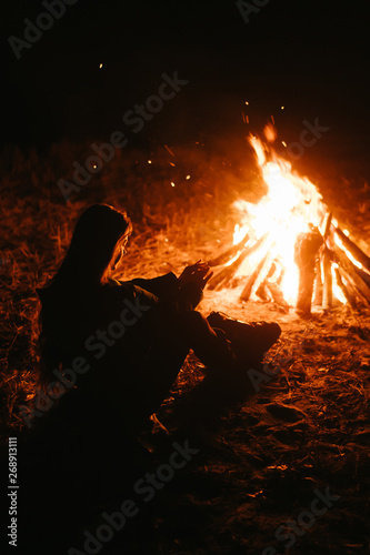 Woman sitting and getting warm near the bonfire in the night forest.
